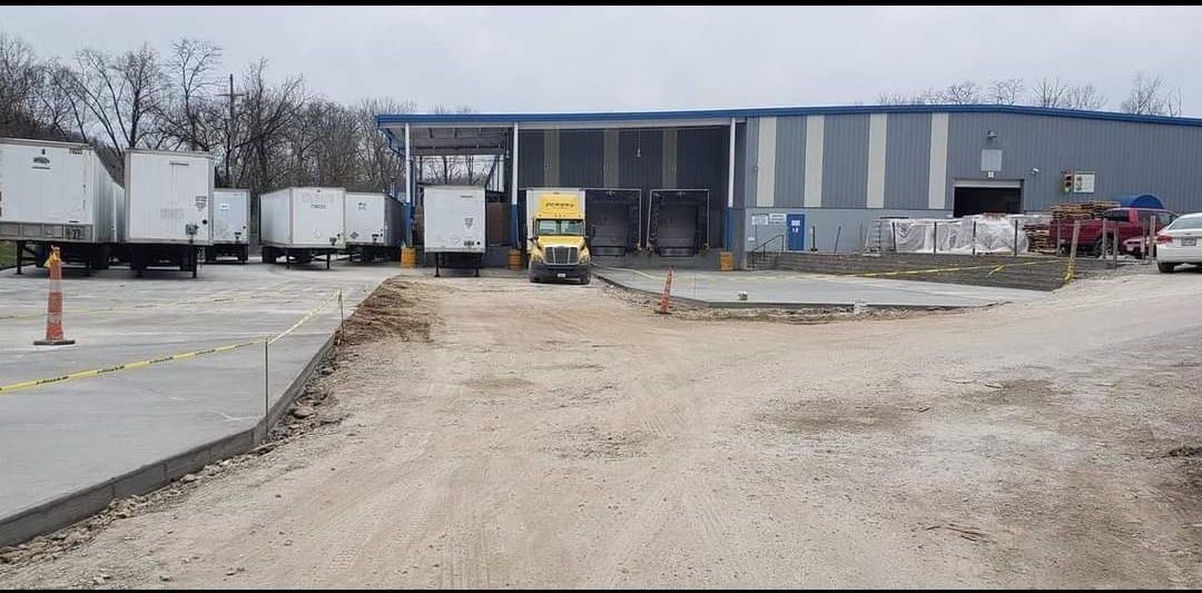 A truck drives toward a warehouse loading dock. Other trailers sit in the lot.