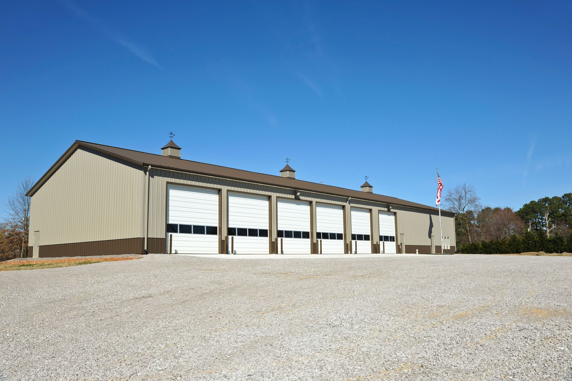 A tan, multi-bay metal garage building under a clear blue sky, situated on a gravel lot with an American flag.