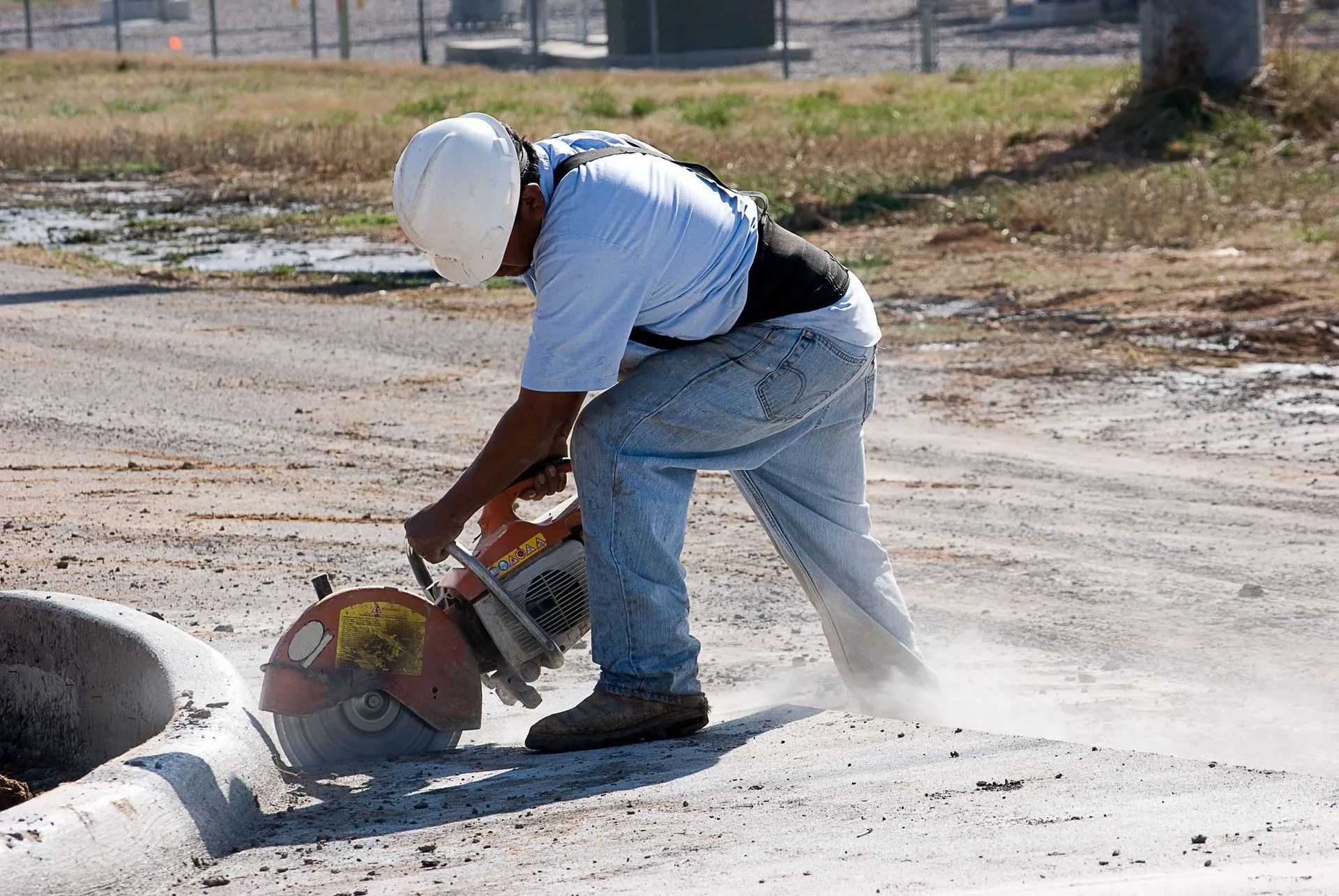 Person in a white hard hat cutting a concrete pipe with a power saw, creating dust. Outdoors, sunny.