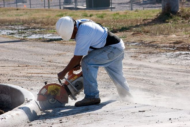 Person in a white hard hat cutting a concrete pipe with a power saw, creating dust. Outdoors, sunny.