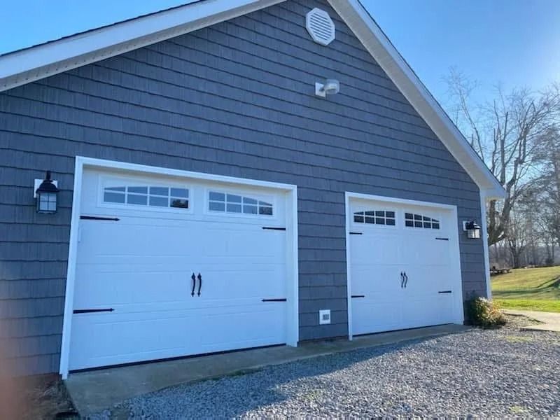 Two white garage doors on a blue-sided building with black hardware and lanterns. Gravel driveway.