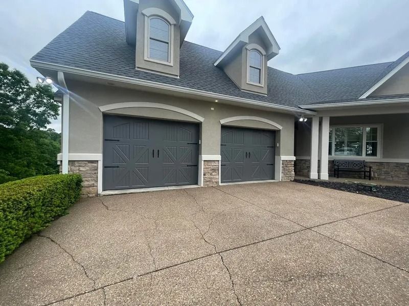 A gray two-car garage with a stone facade, driveway, and dormer windows under a cloudy sky.