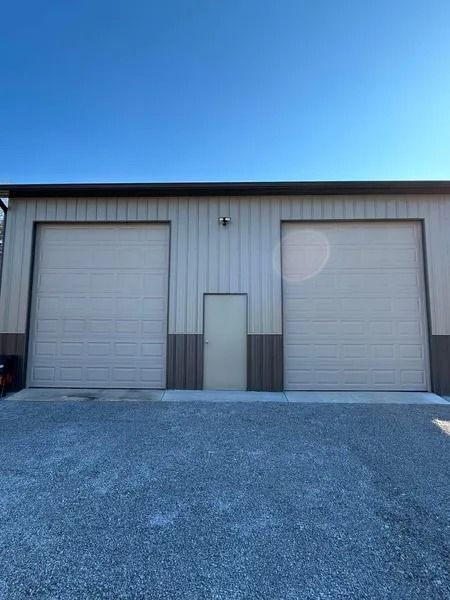 Exterior of a tan metal building with two garage doors and a central door. Gravel ground, clear sky.