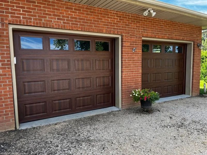Two brown garage doors on a red brick building with a gravel driveway.
