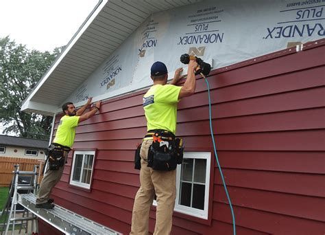 Two workers install red siding on a house, using a nail gun.