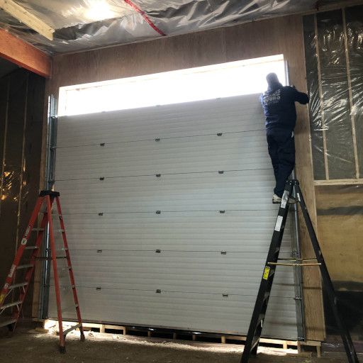 A person installs a white garage door in a building. They are on a ladder.