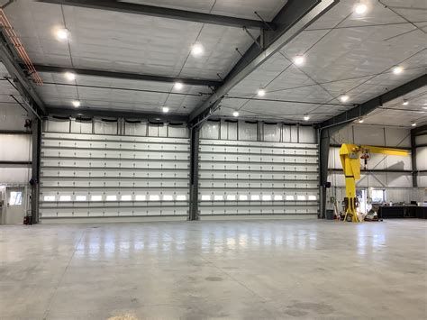 Large industrial warehouse interior with open overhead doors, concrete floor, metal beams, and bright lights.