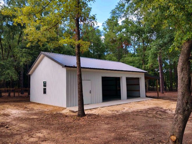 A white metal garage with a dark roof and two open bays, situated in a wooded area on a sandy lot.