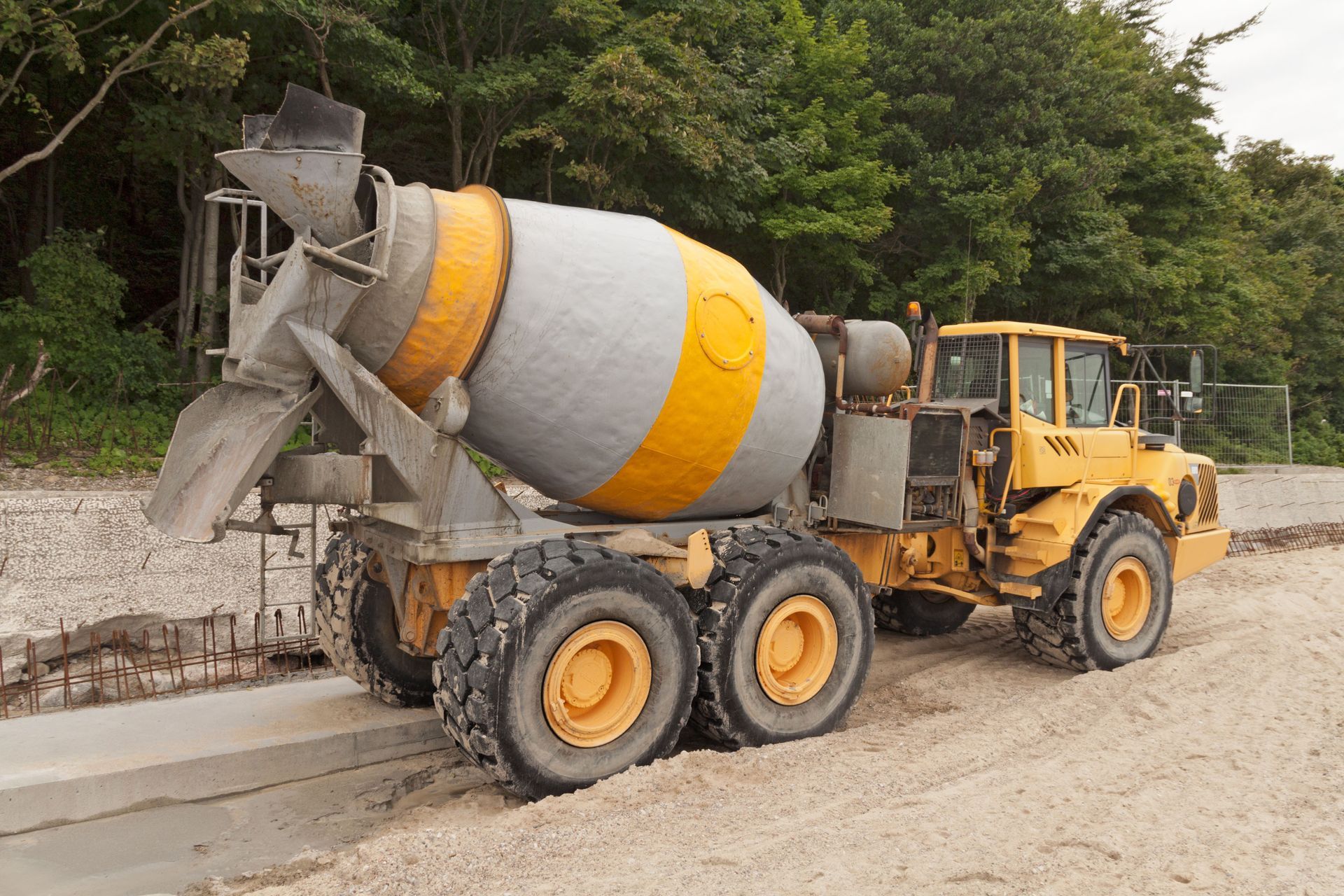 A yellow concrete mixer truck sits on a gravel construction site near a tree line.