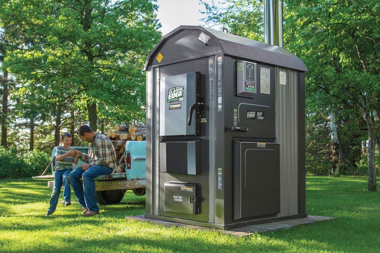 An outdoor wood furnace stands on a grassy lawn near a small utility trailer loaded with logs in a wooded setting.