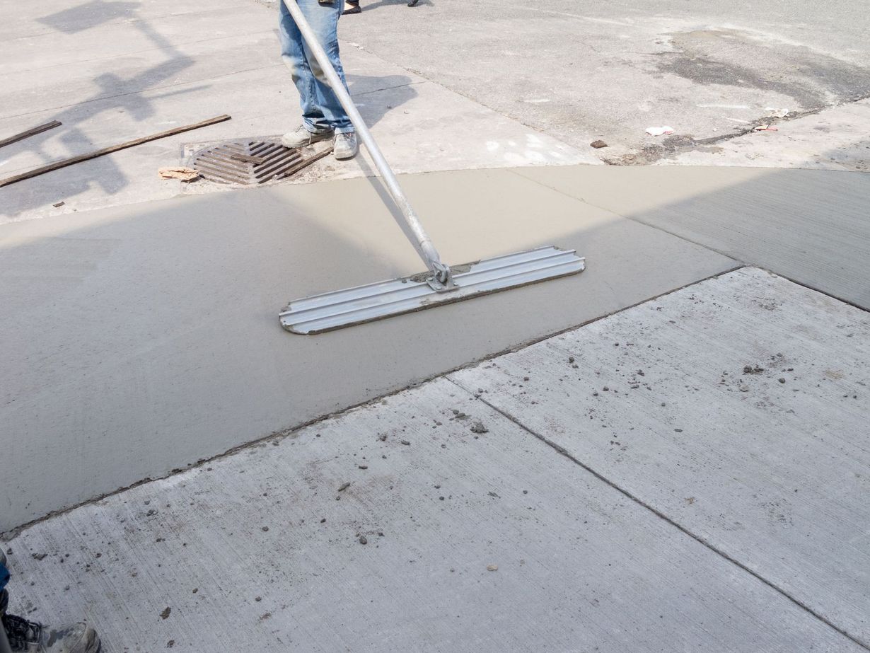 A person uses a long-handled bull float to smooth wet concrete on a construction site.