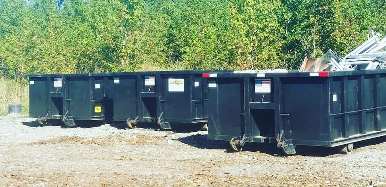 Three black, empty roll-off dumpsters sit in a row on a gravel lot in front of a tree line.