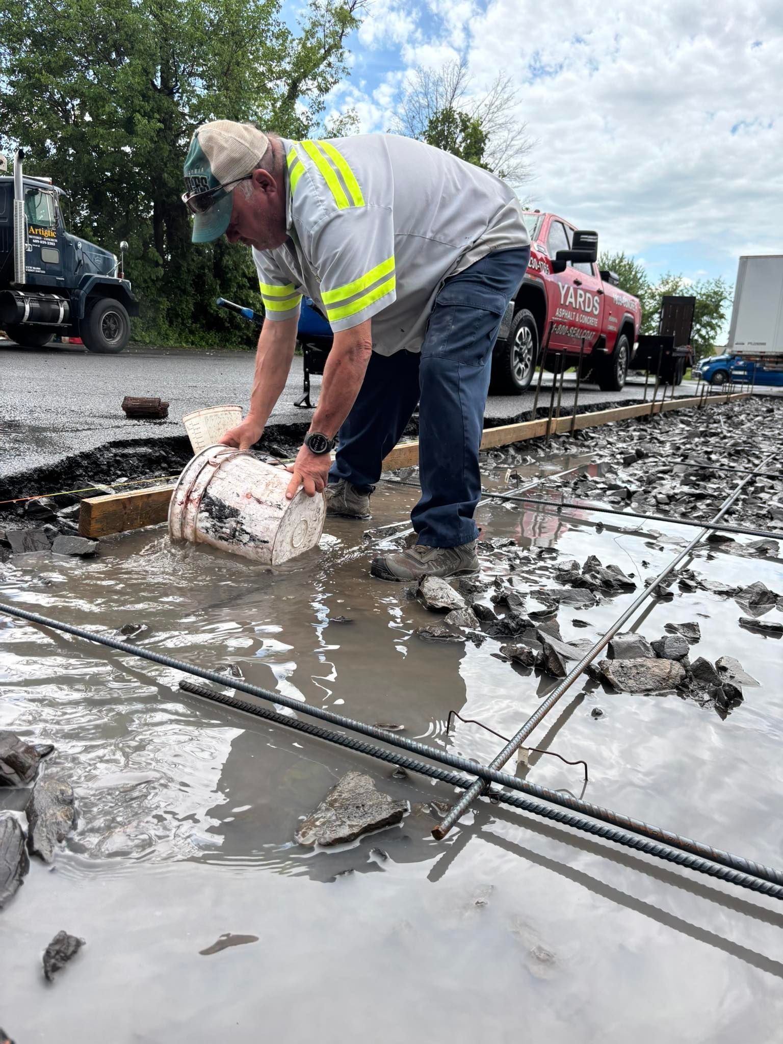 A construction worker in a high-visibility shirt pours a white bucket into a flooded area with rebar and wooden forms.