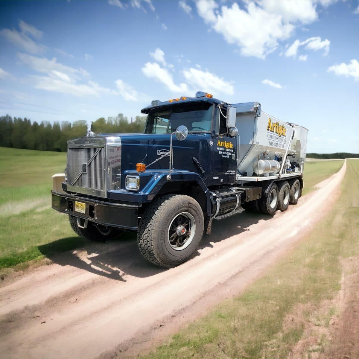 A blue industrial truck with a concrete mixer travels on a dirt road through a grassy field under a sunny blue sky.