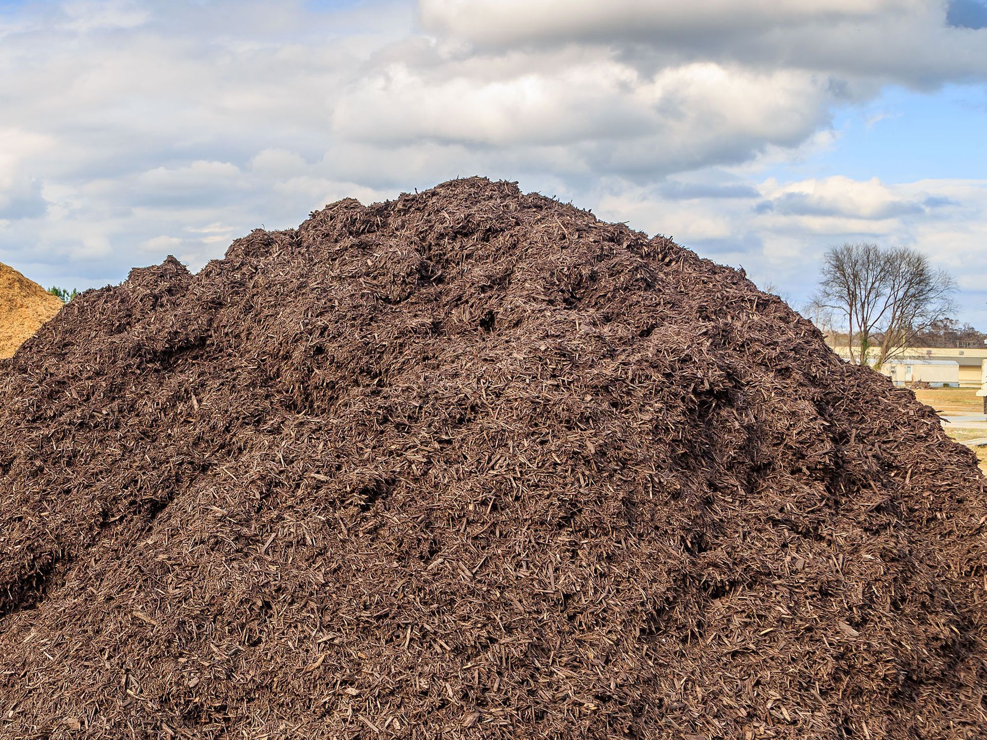 A large, cone-shaped pile of dark brown wood chip mulch outdoors under a blue sky with white clouds.