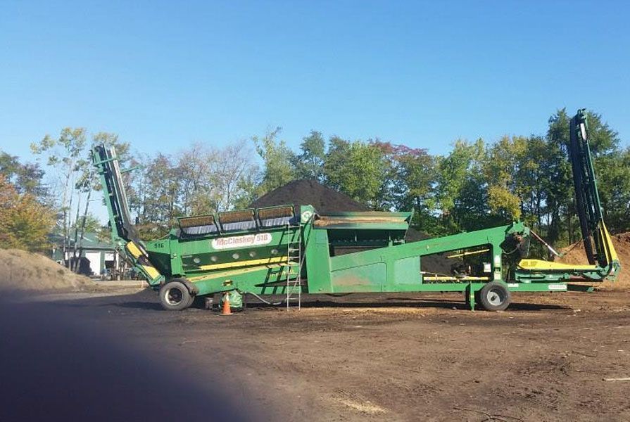 A green industrial trommel screener sits in a dirt clearing, with conveyor belts extended at each end.