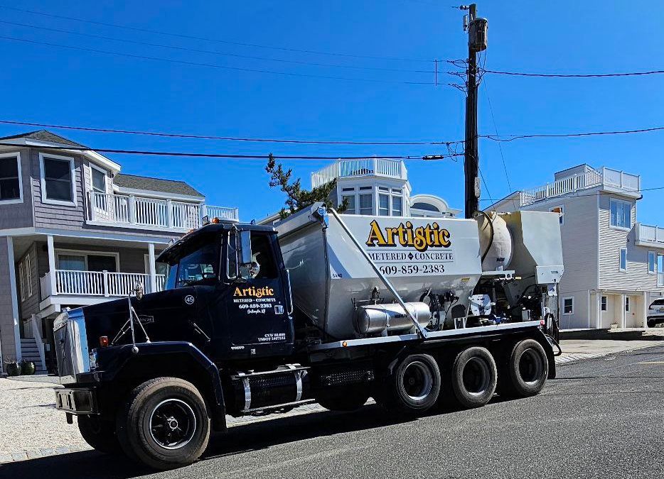 A black Artistic Concrete dump truck is parked on a paved street in front of coastal houses under a clear blue sky.