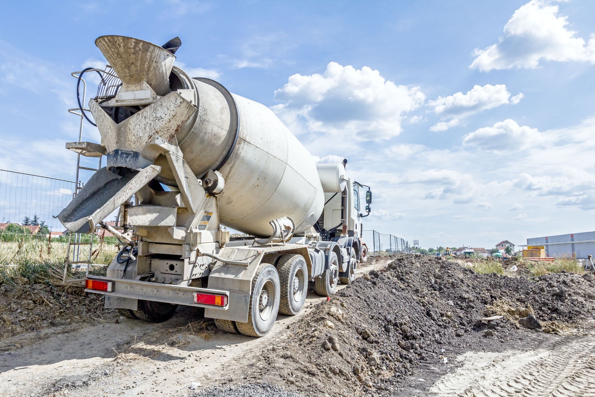 A concrete mixer truck parked on a dirt construction site against a bright, cloudy sky.
