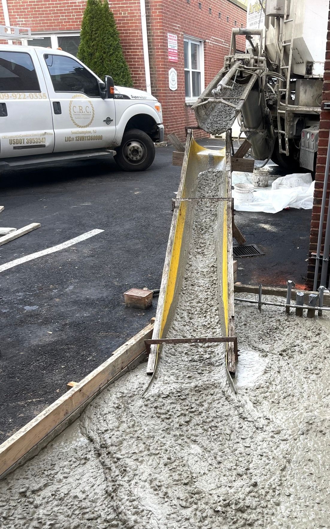 A cement truck pours wet concrete down a yellow chute into a prepared wooden form on a construction site.
