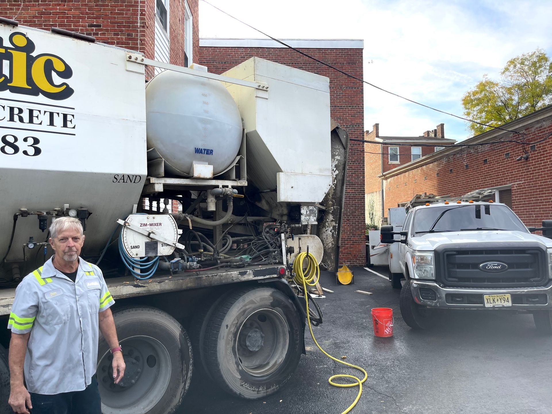 A person in a work shirt stands in front of a concrete mixer truck parked next to a white pickup truck in an alleyway.