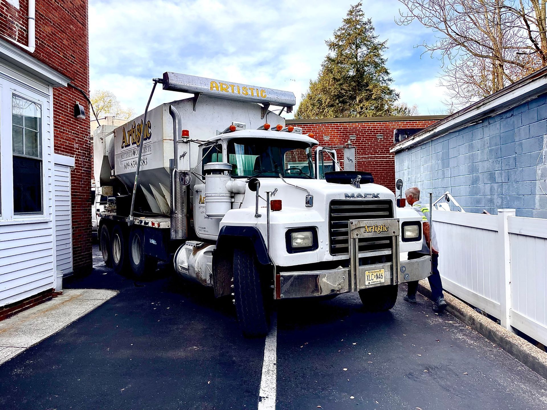 A white heavy-duty utility truck parked in a narrow driveway between a red brick building and a white fence.