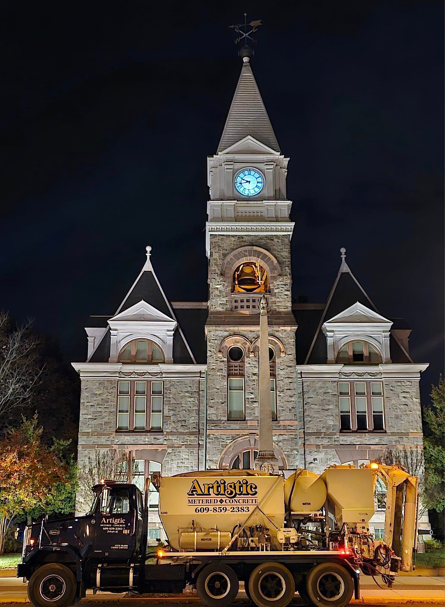 A large, beige construction truck parked at night in front of an illuminated stone clock tower building.