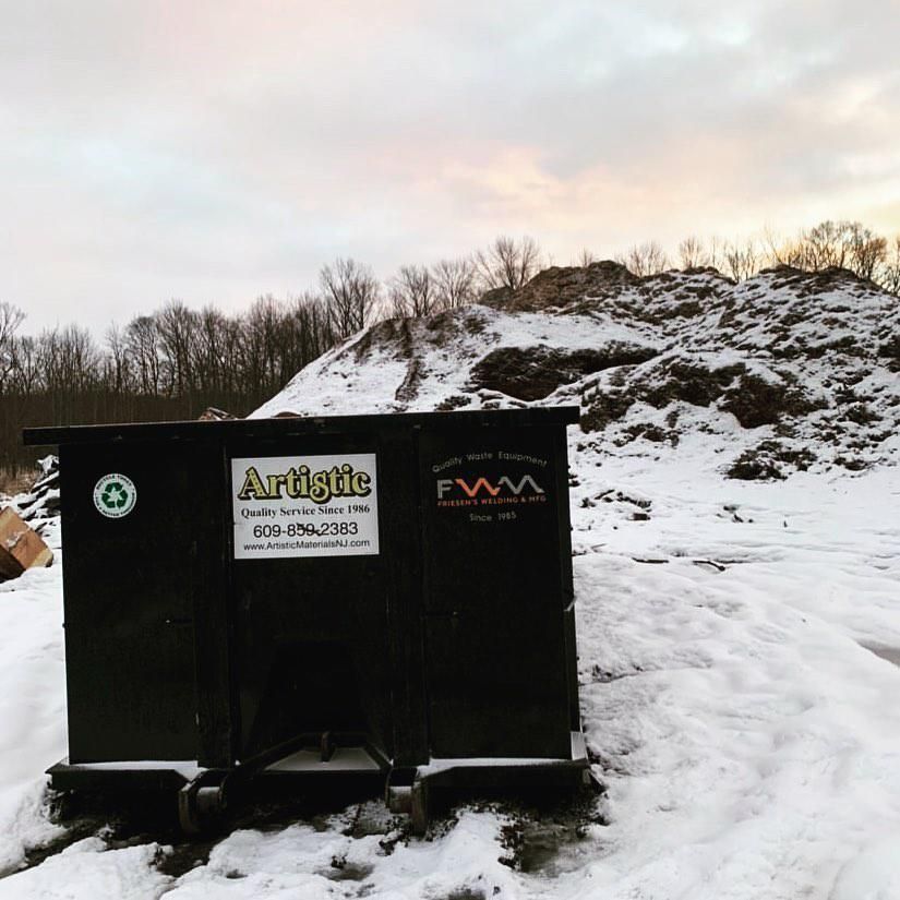 A black dumpster from Artistic Waste Services sits in a snowy field with a large debris pile in the background.