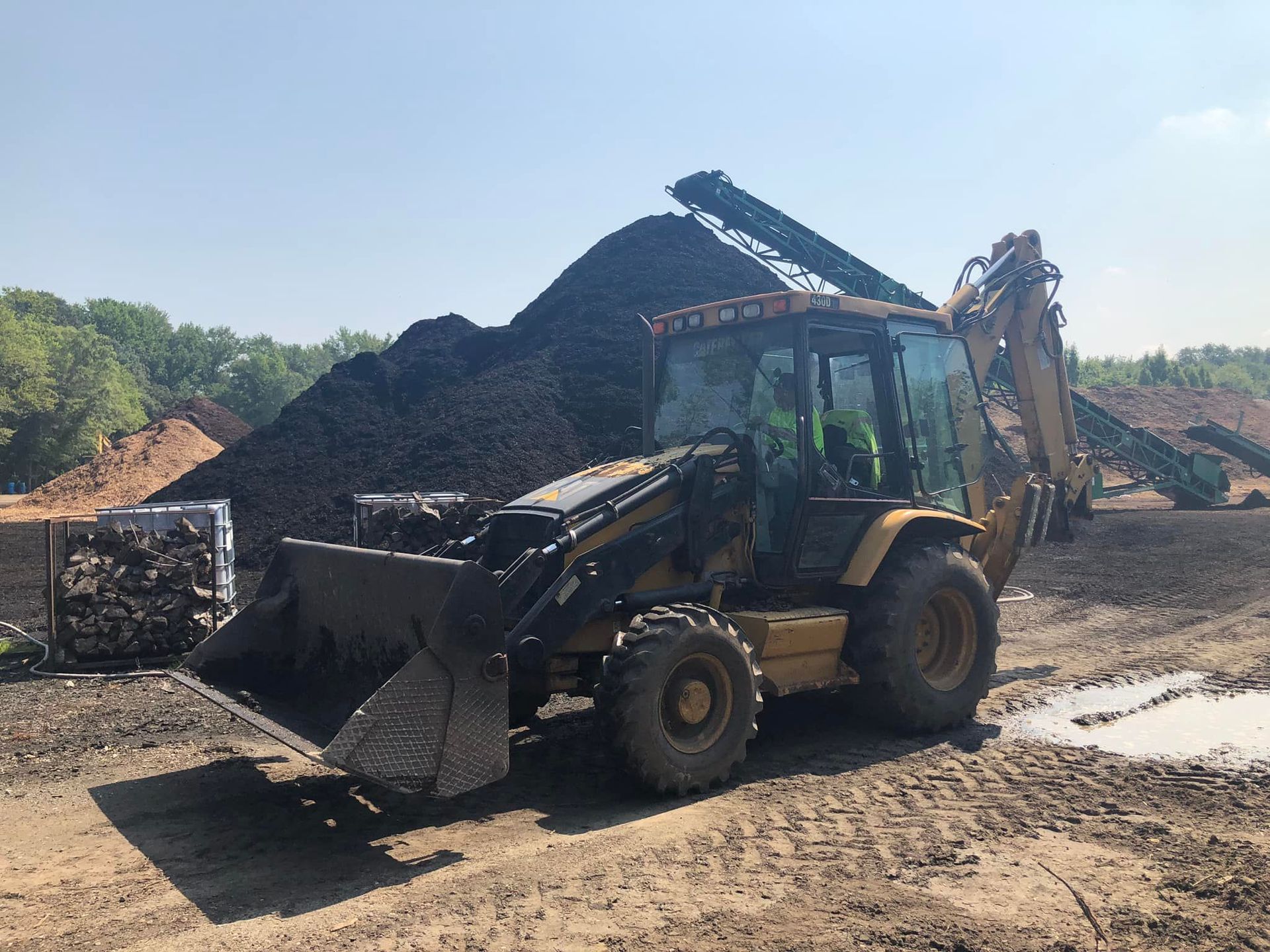 A yellow backhoe loader sits on a muddy work site in front of a large pile of dark organic material and a conveyor belt.