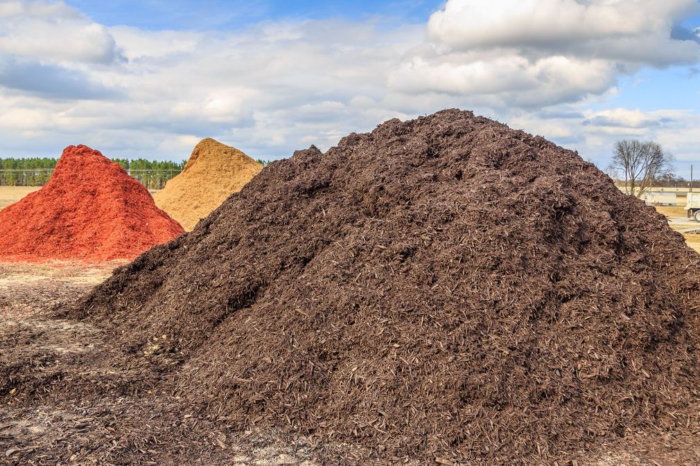 Three large, separate piles of red, gold, and dark brown mulch sit in an open field under a cloudy blue sky.