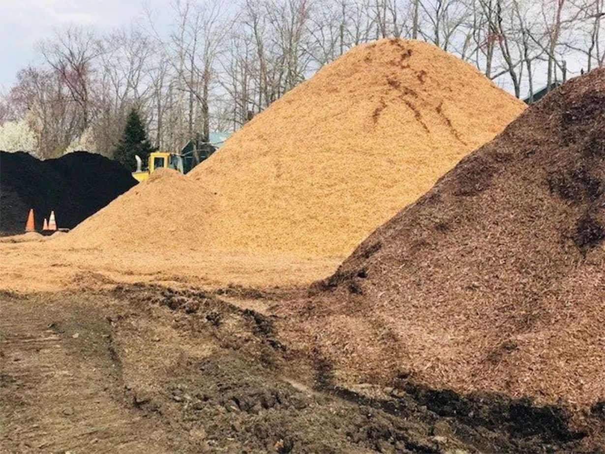 Large piles of light-colored wood chips and dark brown mulch sit in an outdoor yard with trees in the background.
