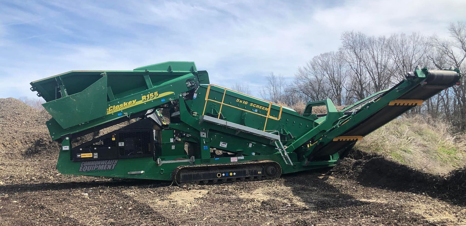 Green track-mounted industrial screening machine sitting on dirt, processing soil outdoors.