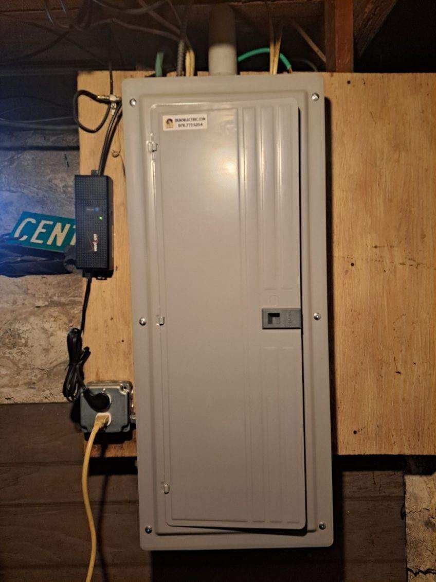 Electrical panel with a closed gray door on a wooden wall, power cord plugged in.