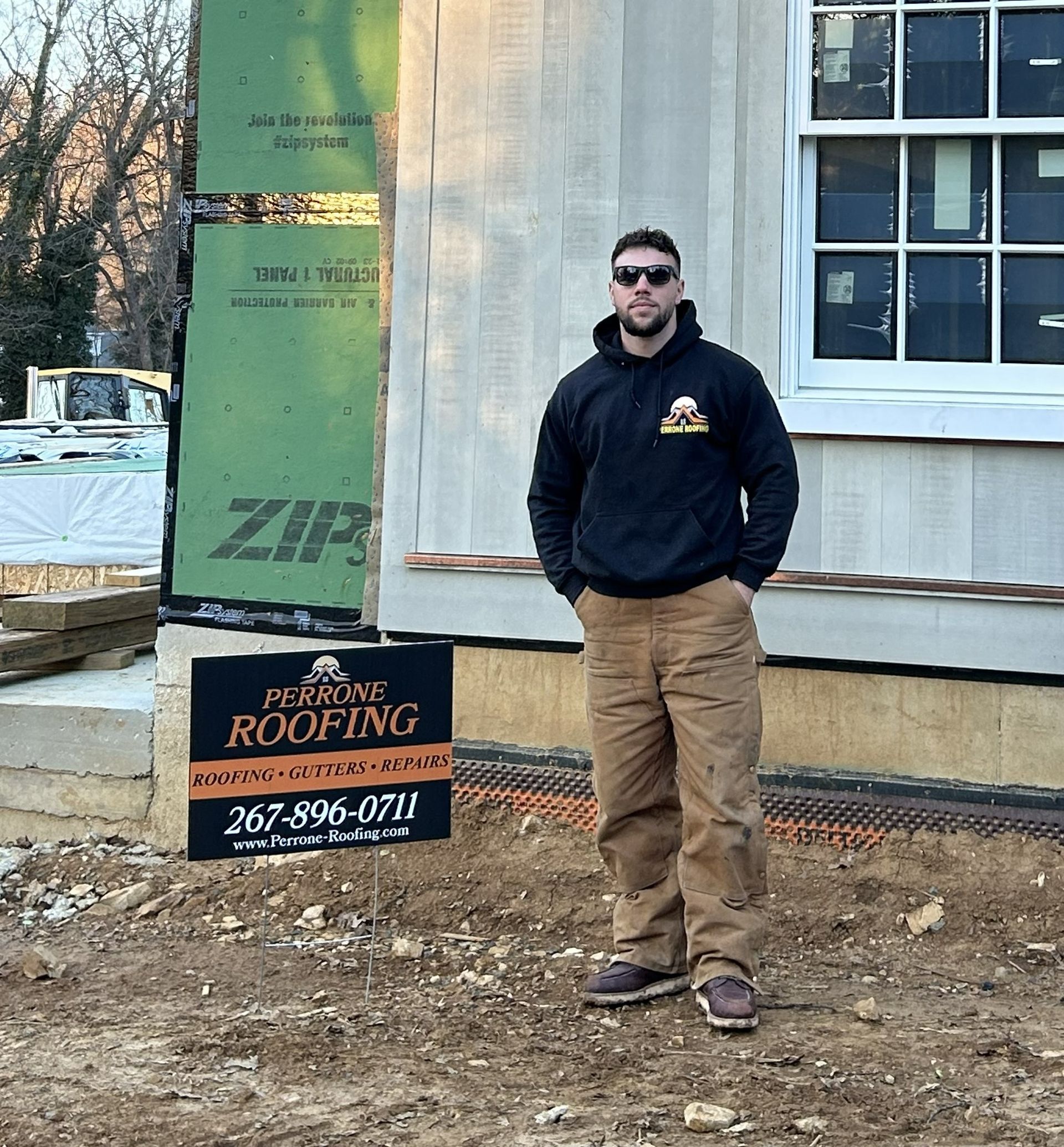 Man standing outside a house under construction beside a roofing sign, wearing a dark hoodie and tan pants.
