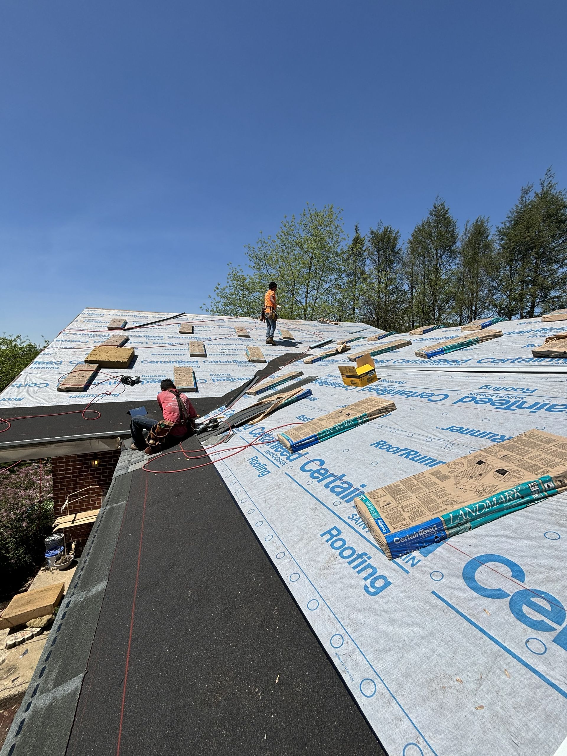 Roofers installing black underlayment on a sloped roof with stacked materials under a blue sky