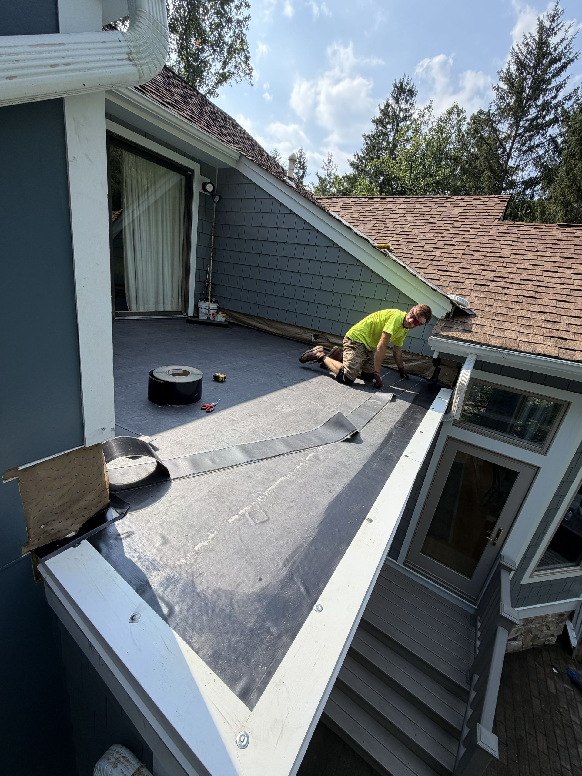 Worker installing roofing on a gray dormer roof beside a house window