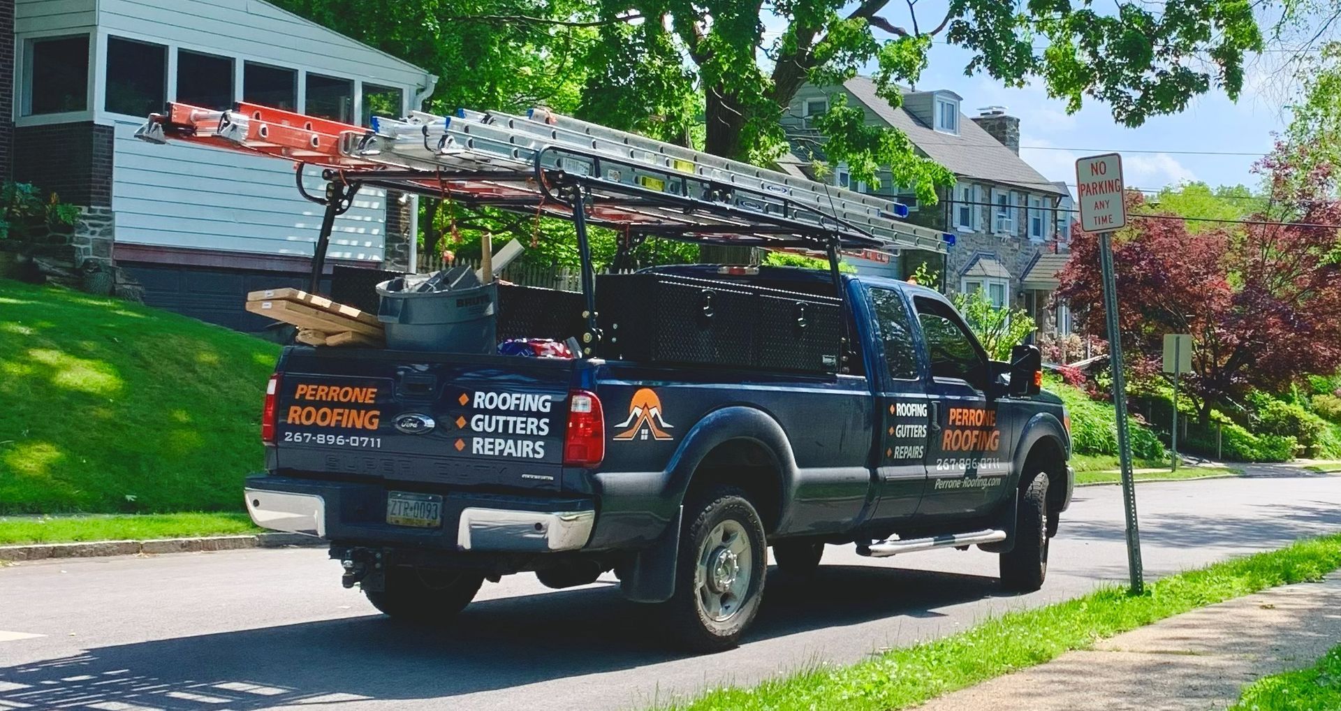 Black utility truck with ladder rack parked on a sunny residential street, houses and green trees behind.