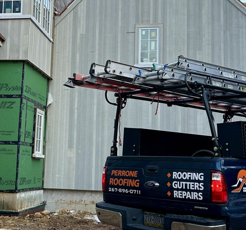 Blue roofing van with ladders parked beside a house under construction and green wrapped materials