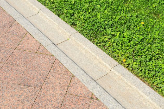 A light stone curb separates a textured, reddish-brown paved path from a patch of vibrant green grass.