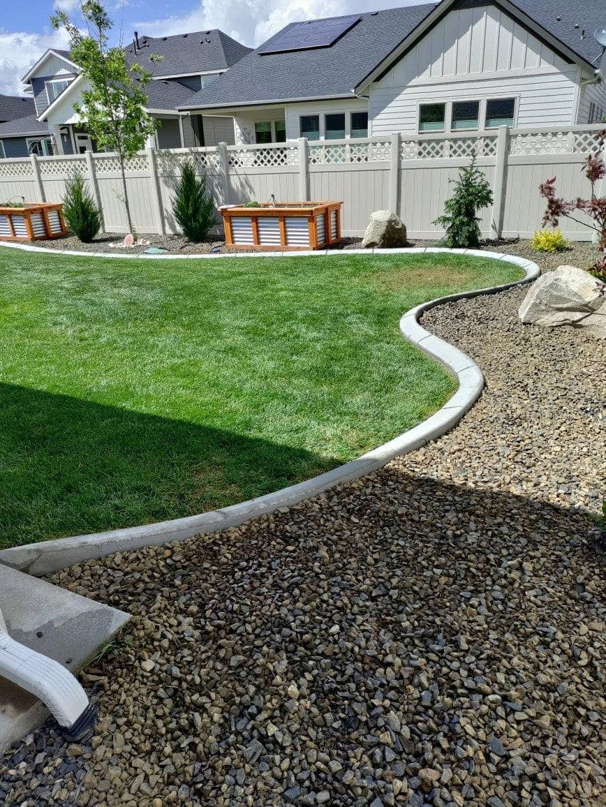 A lush green lawn with a curved concrete border, separated from a dark gravel area, backed by a white fence and houses.