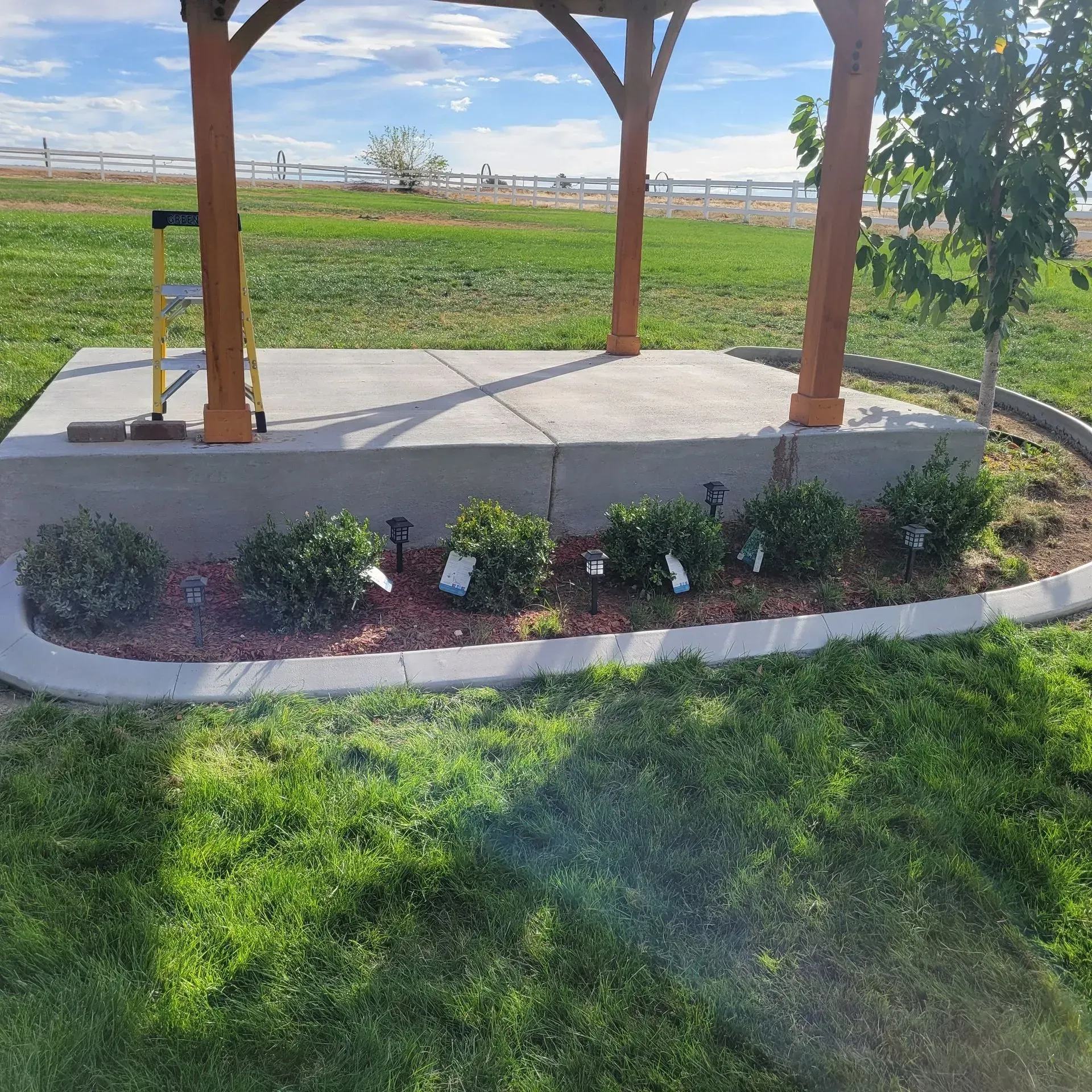 A wooden gazebo structure stands over a concrete platform, bordered by a garden bed with small shrubs and landscaping lights.
