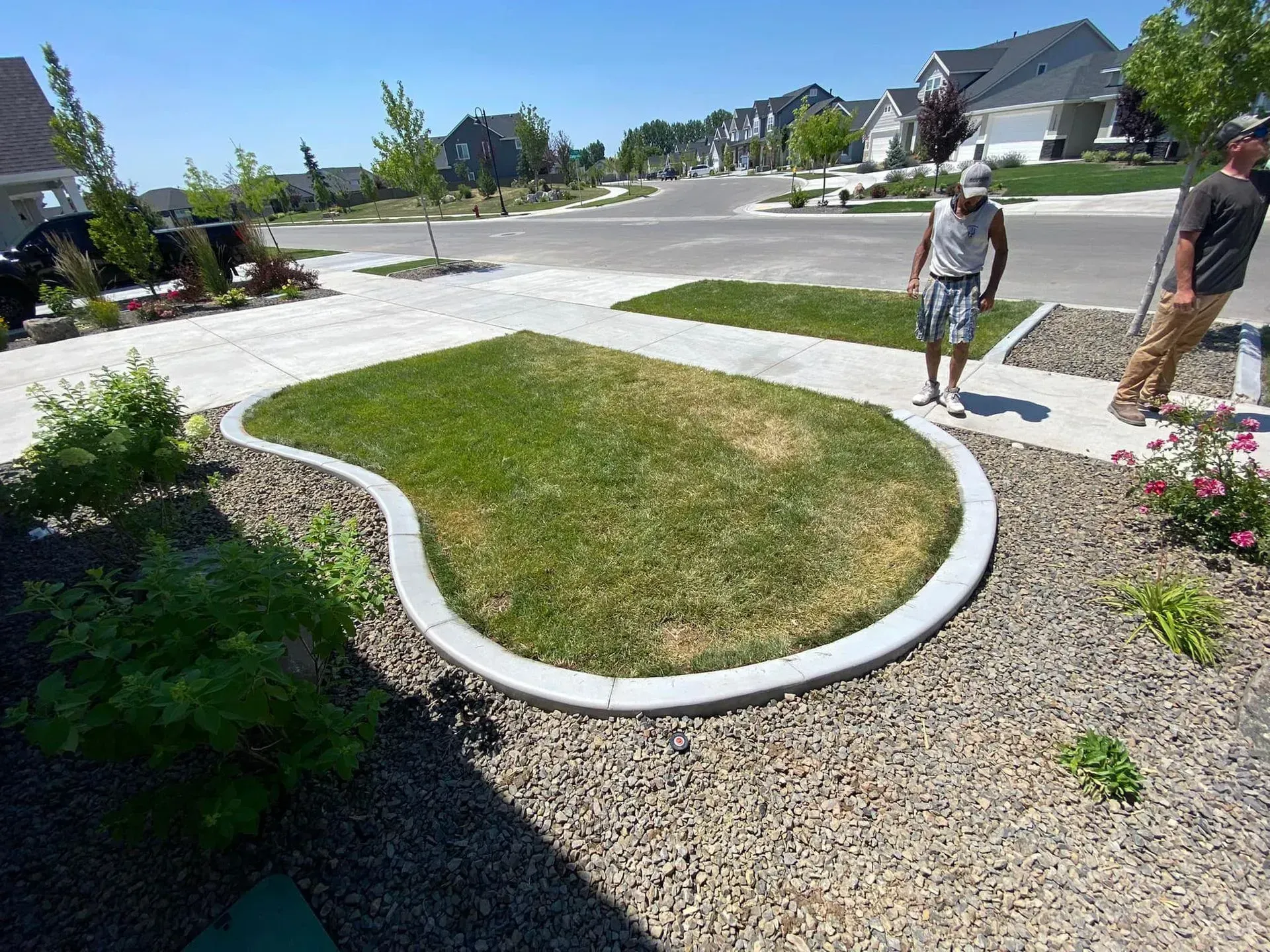 Two people stand on a sidewalk next to a small, kidney-shaped grass patch surrounded by decorative stone in a suburban yard.