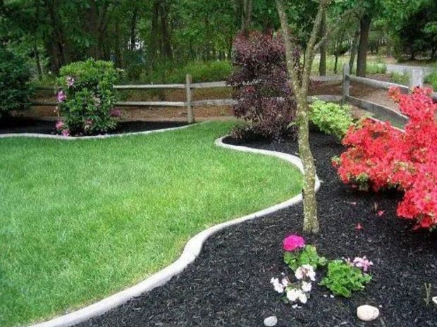A manicured lawn with a curved concrete border separating it from a black-mulched garden bed with shrubs and red flowers.