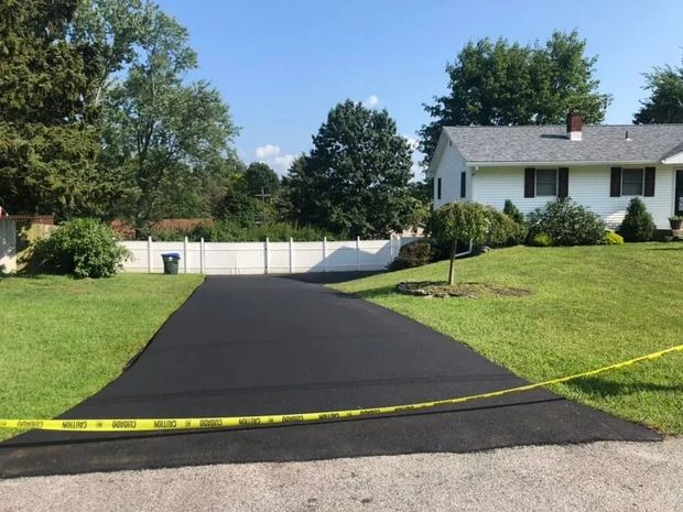A freshly paved black asphalt driveway leads to a white house with a lawn and a white fence under a clear blue sky.