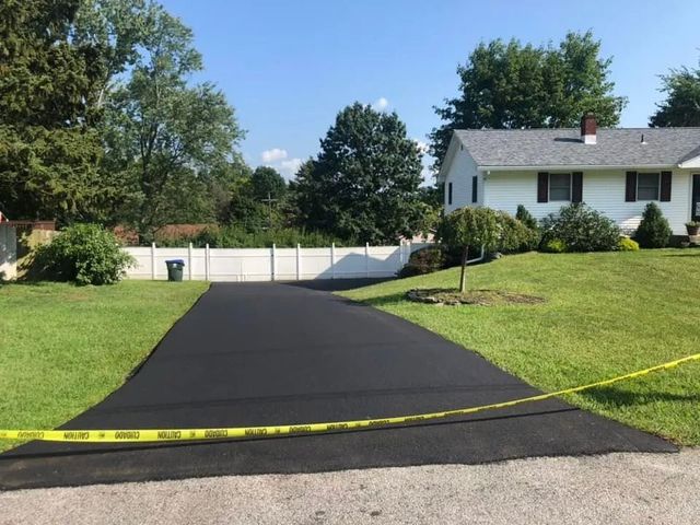 A freshly paved black asphalt driveway leads to a white house with a lawn and a white fence under a clear blue sky.