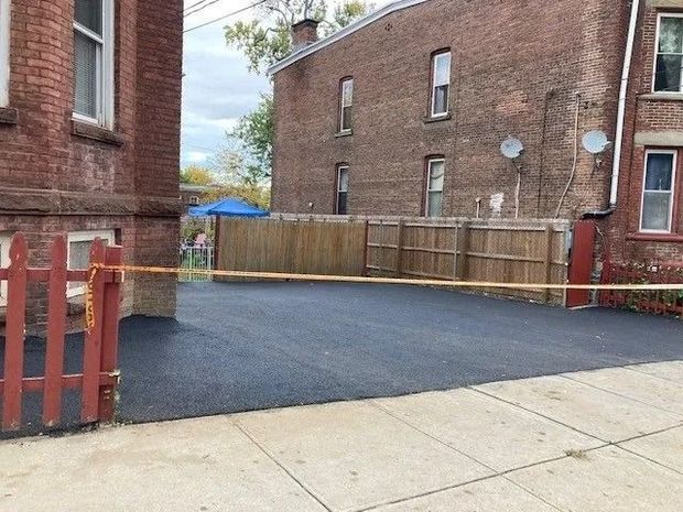 A newly paved asphalt driveway in front of brick buildings, bordered by a wooden fence and a red gate with caution tape.