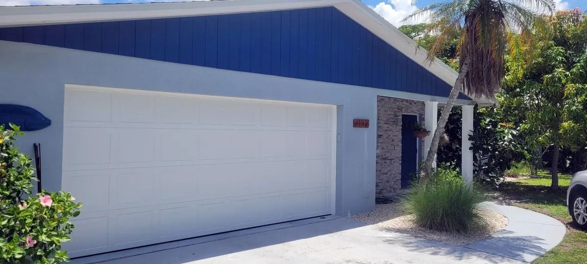 A light blue house with a white garage door and dark blue roof. There is a small bush and some trees.