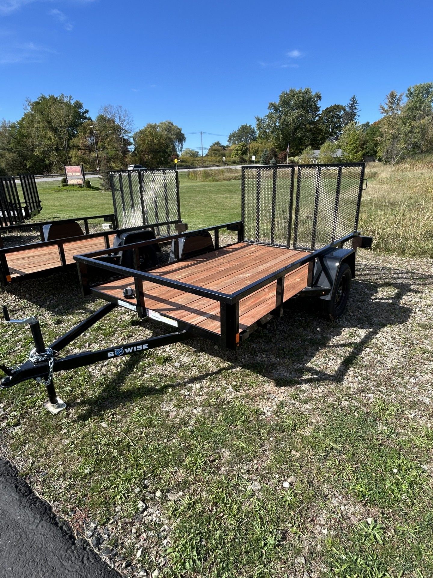 Two black utility trailers with mesh sides and wood floors on grass under a blue sky.