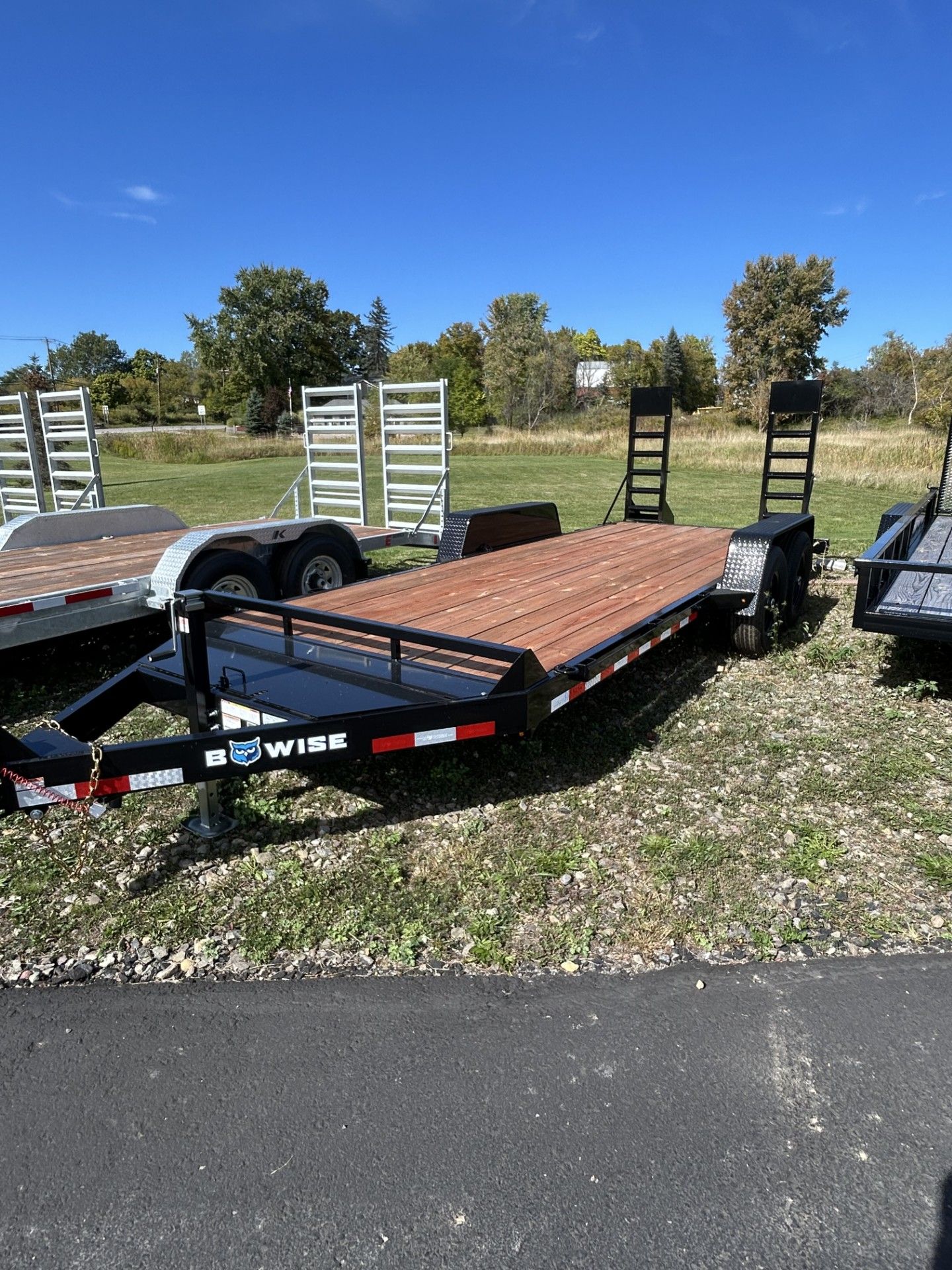 Black and brown low-profile trailer with wooden deck on grass against a blue sky.