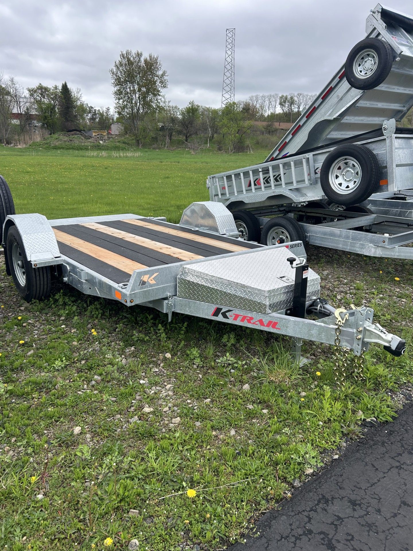 Car trailer on grass with diamond plate deck. Another trailer is in the background. Cloudy sky.