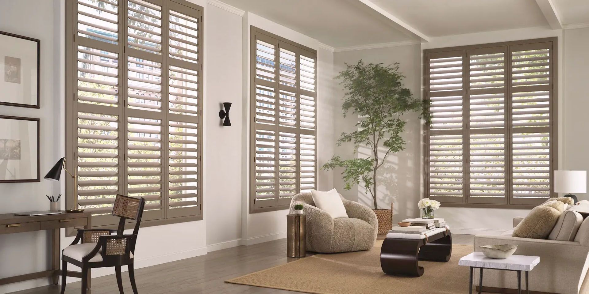 Bright living room with shuttered windows, beige sofas, a chair, coffee table, and potted plant